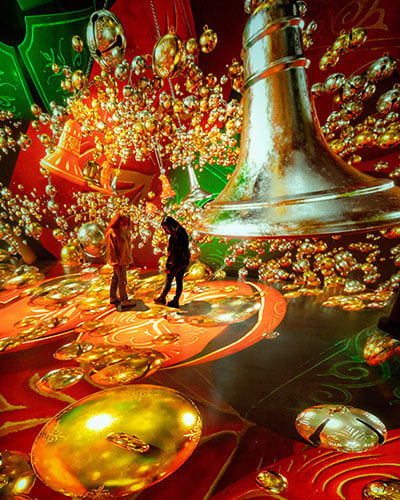 Two people look at images of gold bells with a green and red background that are projection mapped onto a wall and floor.