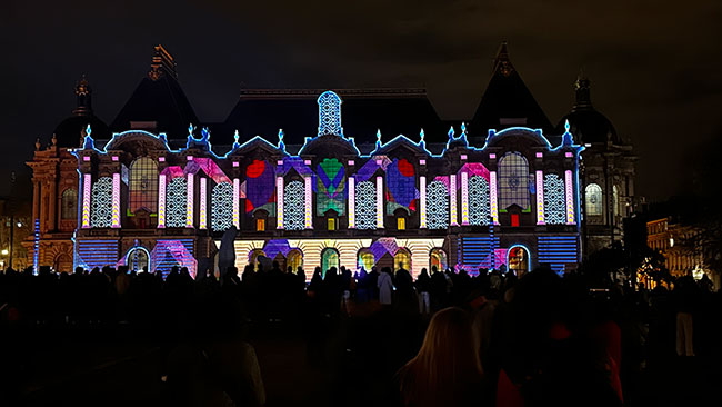 The façade of a building is projection mapped with various colors and patterns. 