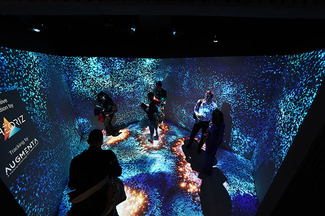 People in a room with walls and floor projection mapped with bright blue and green dots. 