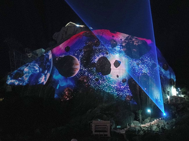A tall rock face is projection mapped with images of planets. 