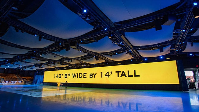 A large yellow LED video wall stretches across an empty hall with the words 143 feet 8 inches wide by 14 feet tall on it. 