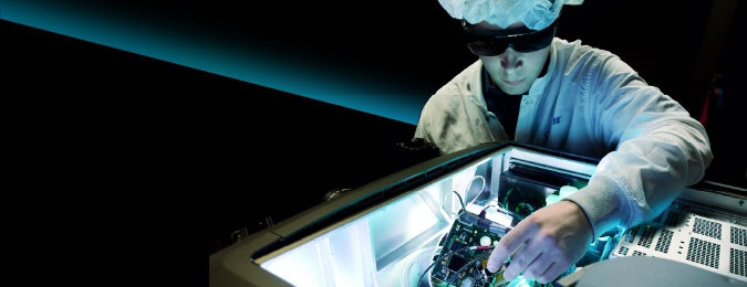 A person wearing safety glasses, a lab coat and hairnet looking at the inside of a projector