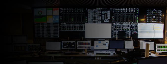 A person sitting in a control room with lots of computer monitors and large video walls showing graphs and data.