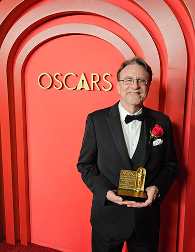 Man dressed in a tuxedo holding a gold award standing in front of a red wall with the word “Oscars” on it. 