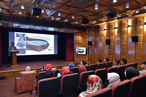 People sitting in seats inside a small theatre, watching a person give a presentation from the stage in front of a screen with a projection on it