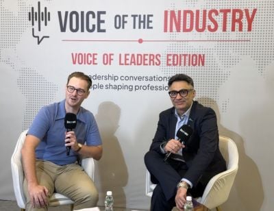 Two people with microphones sitting in white chairs in front of a 