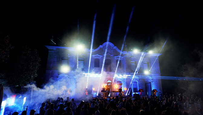 A crowd of people at a music festival, with projection mapping illuminating a building. Photo credit @ADCL Audiovisuais