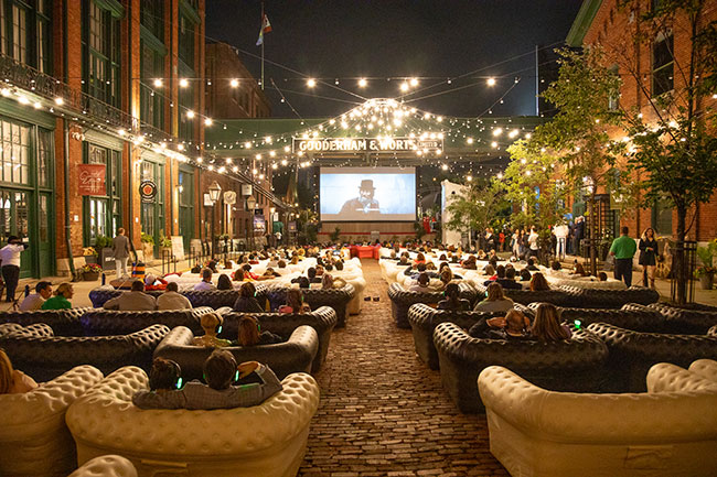     An audience sits in couches placed along a cobblestone street watching a film.  