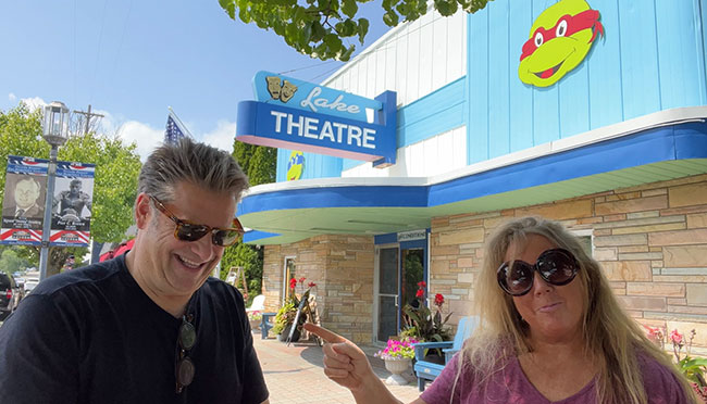A woman is pointing to a man beside her, and they are standing in front of a building with beige stone and blue and white siding with a sign that reads “Lake Theatre”.