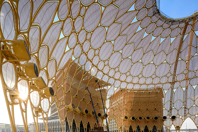Large curved wall featuring trellis design with opaque fabric stretched between the pieces. Buildings visible in the distance. 
