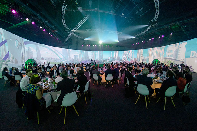 Professionally dressed people sitting at round dining tables with a 360 degree curved screen in the background projecting an urban setting