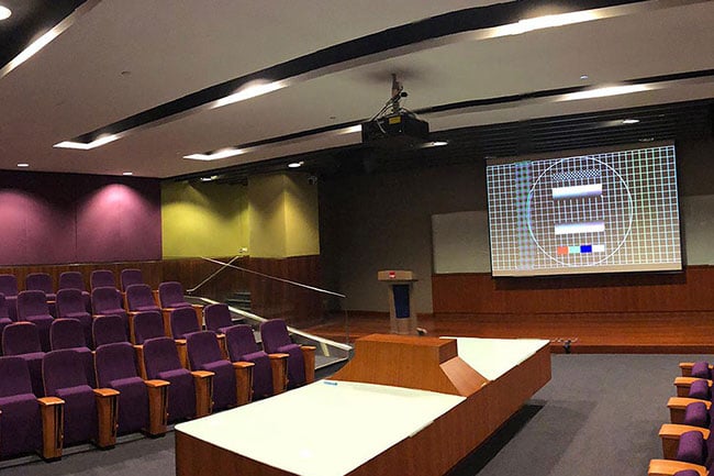 a small lecture room with rows of seats on the left and right and a Christie projector hanging from the ceiling projecting onto a screen