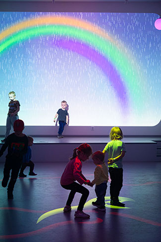A rainbow is projected onto a wall, with children playing in the foreground.