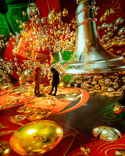 Two people look at images of gold bells with a green and red background that are projection mapped onto a wall and floor.
