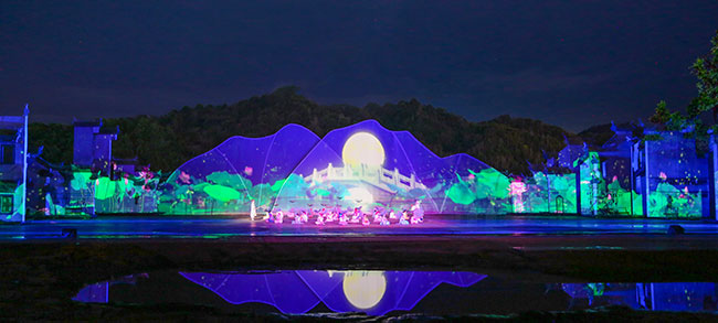 Performers on a large stage with digital projections against real-life scenery in the background.