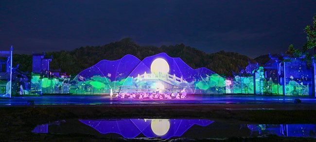 Performers on a large stage with digital projections against real-life scenery in the background.