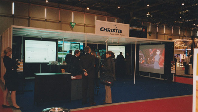 A tradeshow booth with a white ceiling, white support columns, and blue carpet. People are inside and screens with images projected onto them cover the interior walls.