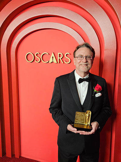 Man dressed in a tuxedo holding a gold award standing in front of a red wall with the word “Oscars” on it.