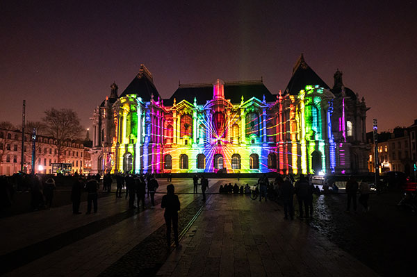 A large historic-looking building is projection mapped in bright, vertical stripes of color. 