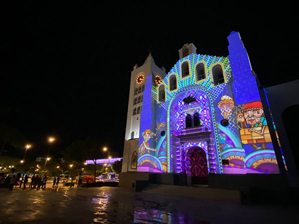 A church is projection-mapped in bright blue, green, and purple with animated images of people.  