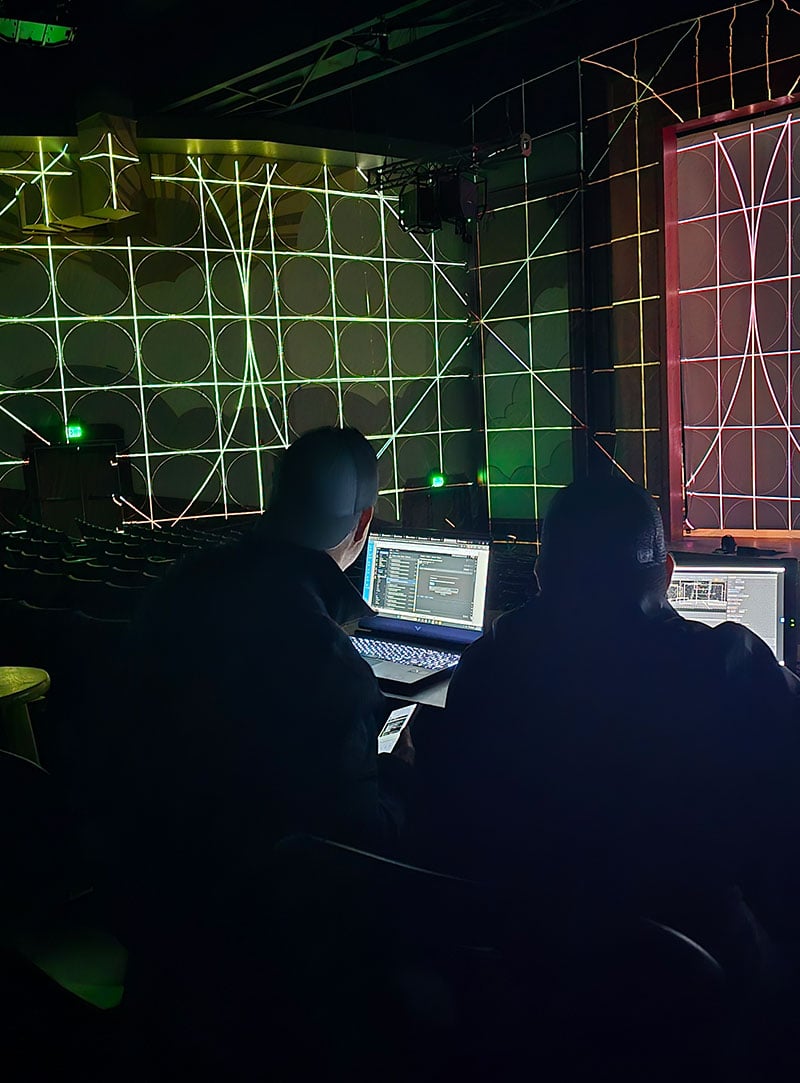 Two people sit at a desk with computers in front of walls projection mapped with a test pattern.