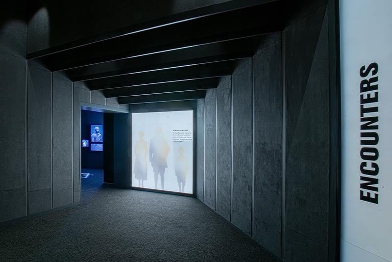 Hallway with gray carpet and walls with the text “Encounters” on one side, and a poster of silhouettes of three people at the end.