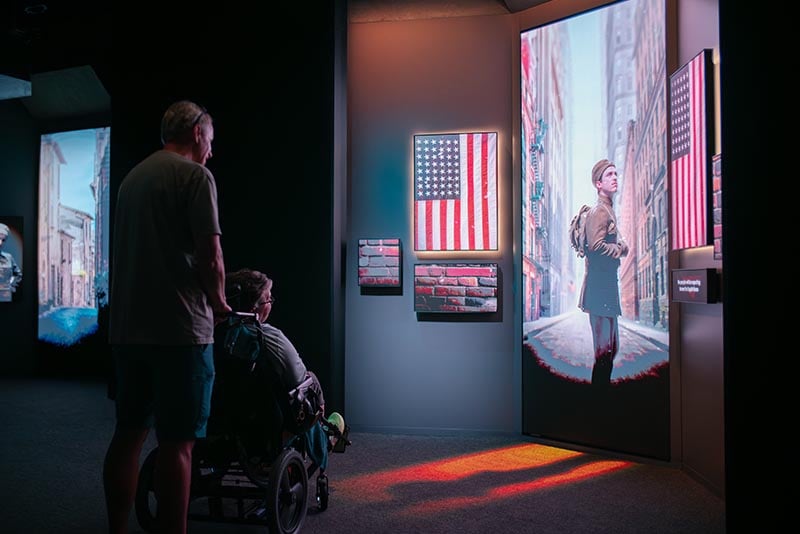 A person standing and a person in a wheelchair look at the image of a man shown on an LED display.