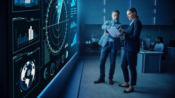 2 professionals standing in front of a large video wall in a bust control room