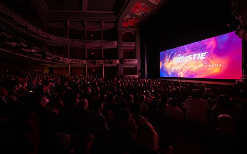 An audience in a large movie theatre facing a screen with the Christie logo on it