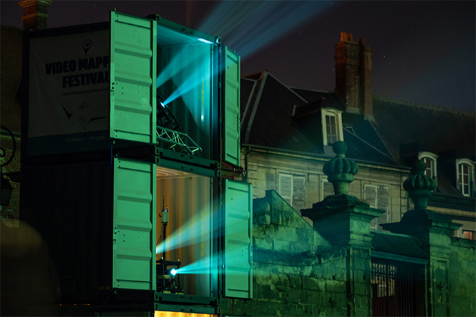 Projectors shine out of two shipping containers stacked on top of one another at night. 