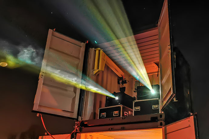 Two projectors shine out of a shipping container at night.