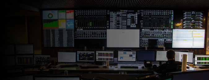 A person sitting in a control room with lots of computer monitors and large video walls showing graphs and data.